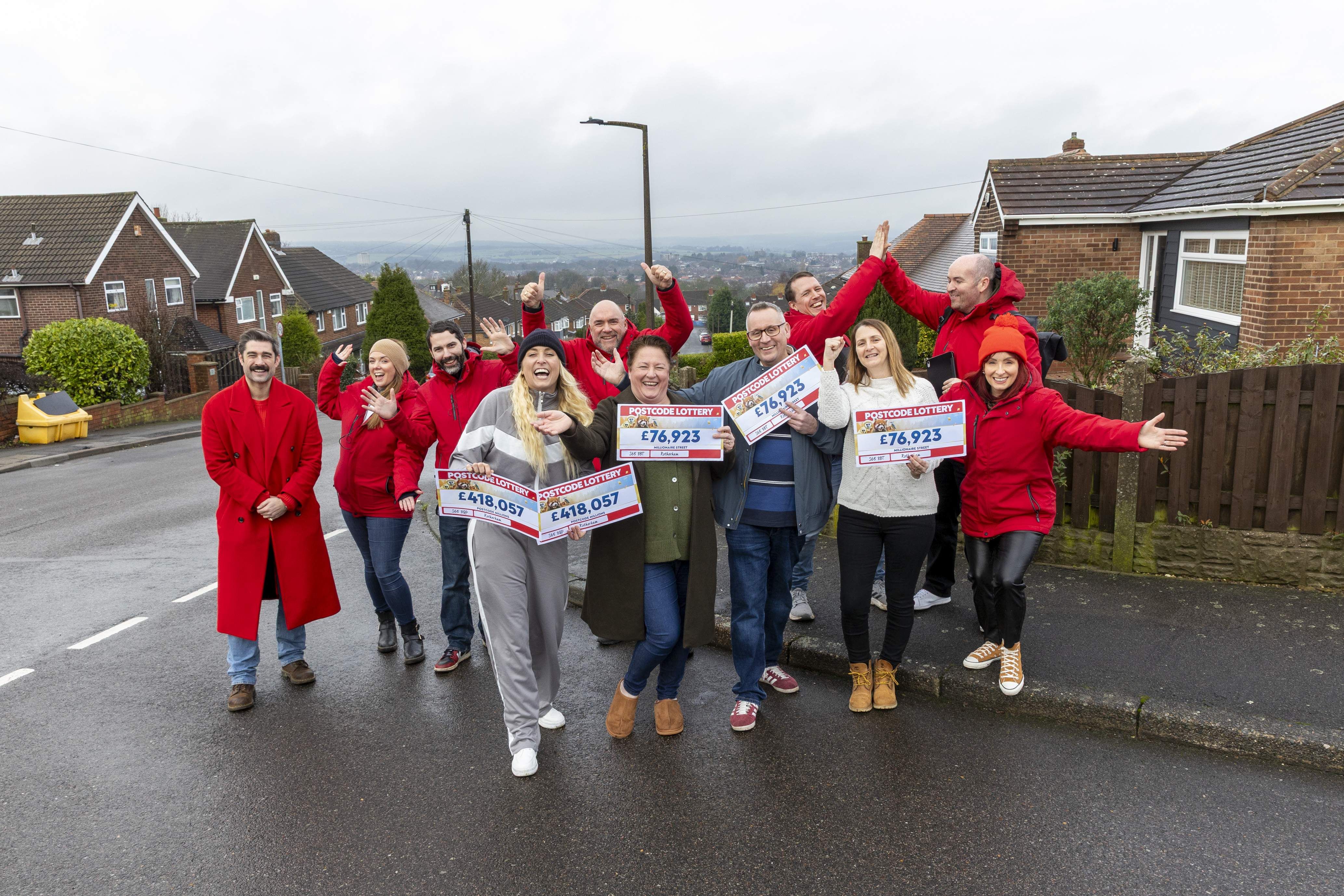 A group of people on a street celebrates, holding large checks from a postcode lottery. Houses and a cloudy sky are in the background.