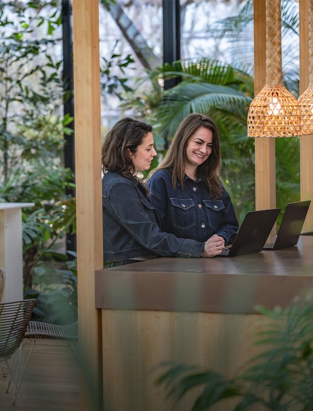 Two women in denim shirts work on laptops at a wooden counter in a lush, green outdoor setting with hanging woven lamps.