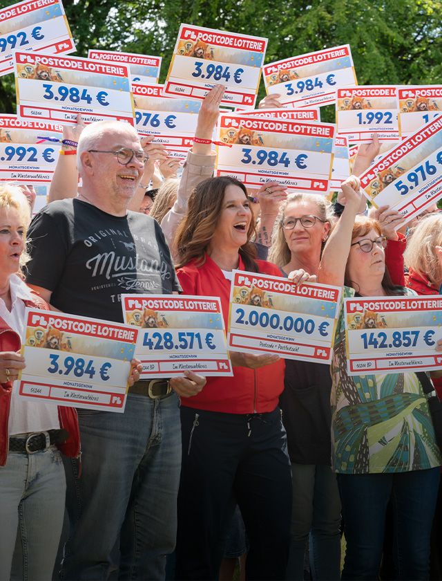 A jubilant group of people holding Postcode Lottery checks, celebrating their winnings outdoors on a sunny day.