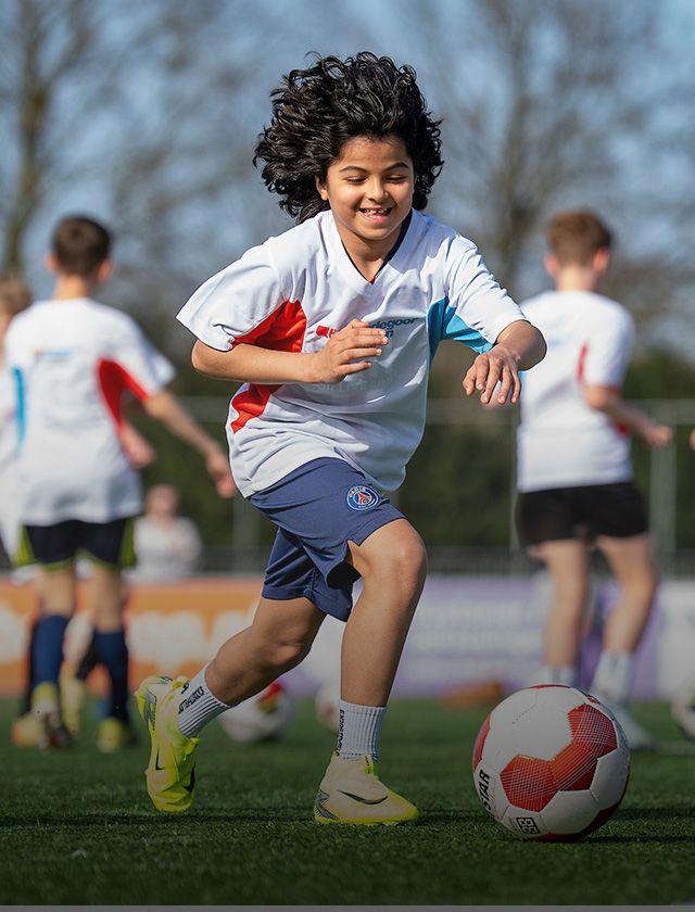 A smiling child with curly hair runs joyfully on a soccer field, wearing a sports uniform, with other kids in similar outfits in the background.