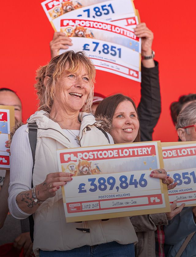 A group of smiling people holding large Postcode Lottery checks with varying amounts against a bright red background.